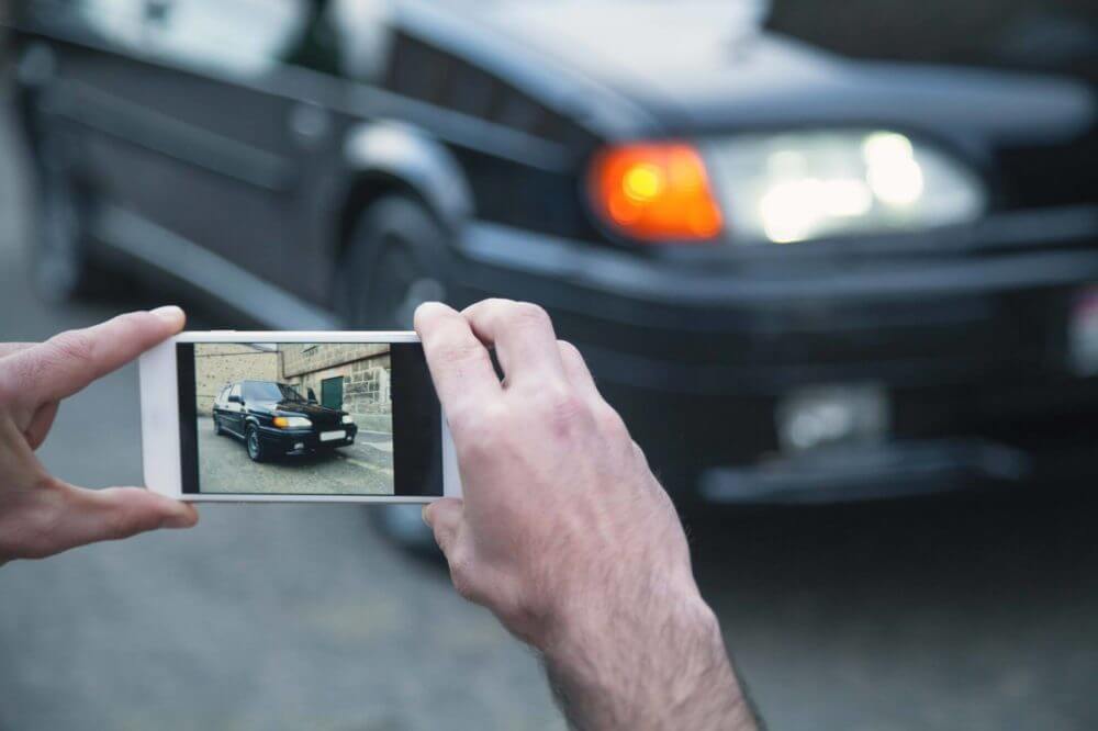  Man is holding his mobile phone with both hands and taking pictures of its black automobile. 