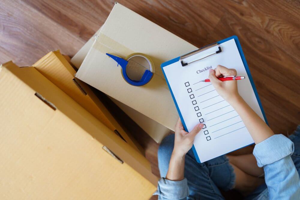 Person writing a checklist surrounded by boxes for cross-country moving