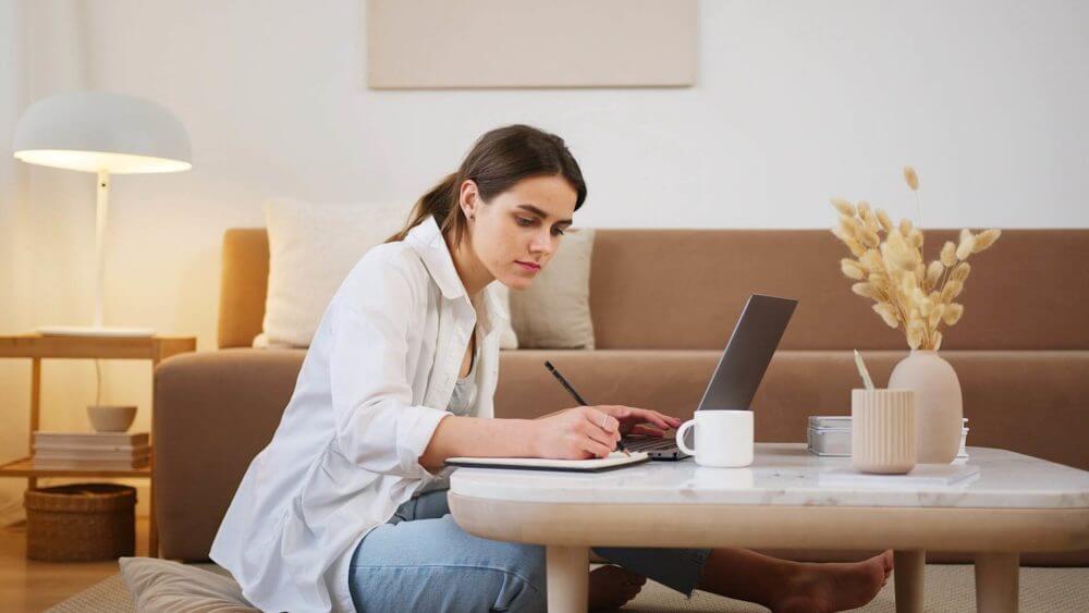 Girl is sitting in her living room at the coffee table and research the internet on a laptop while taking notes and drinking coffee