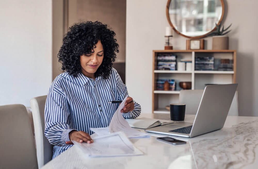 a woman is sitting at her desk, browsing the internet on her laptop, and looking at papers 