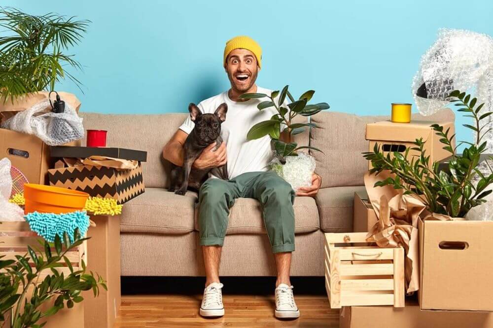 a man holding a small dog and a plant, sitting on a couch surrounded by boxes
