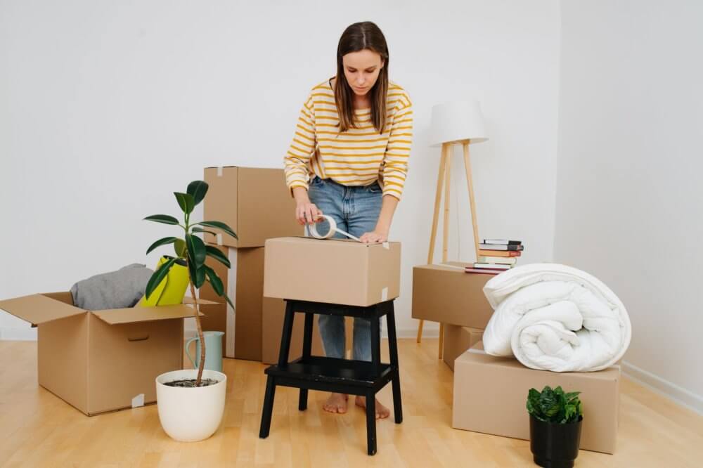 A girl taping the box on the stool, surrounded by boxes for long-distance moving