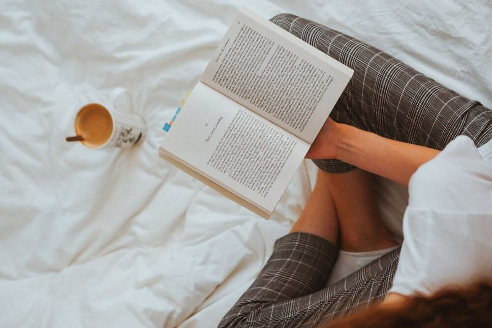 A person in grey pants, reading a book on the bed, a coffee mug on the bed