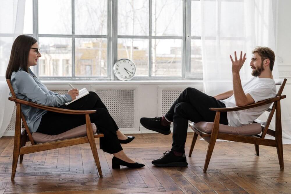 A man in a chair talking to a female therapist sitting across from him 