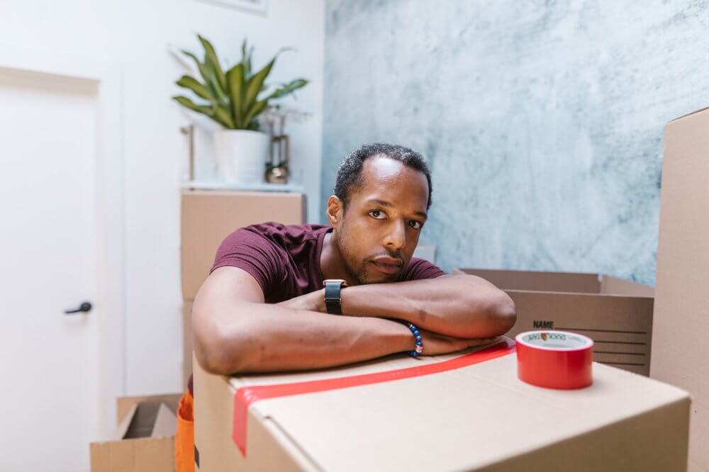 A sad man leaning on the box, other boxes, and a plant behind him
