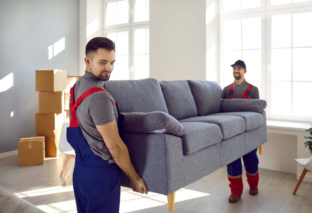 Two professional relocation service workers in overalls moving a sofa