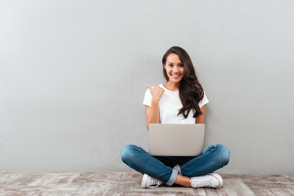 Happy smiling woman working on laptop computer while sitting on the floor with legs crossed and pointing finger away isolated over gray background